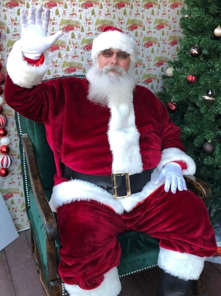 Santa Tom smiling and waving near a Christmas tree.