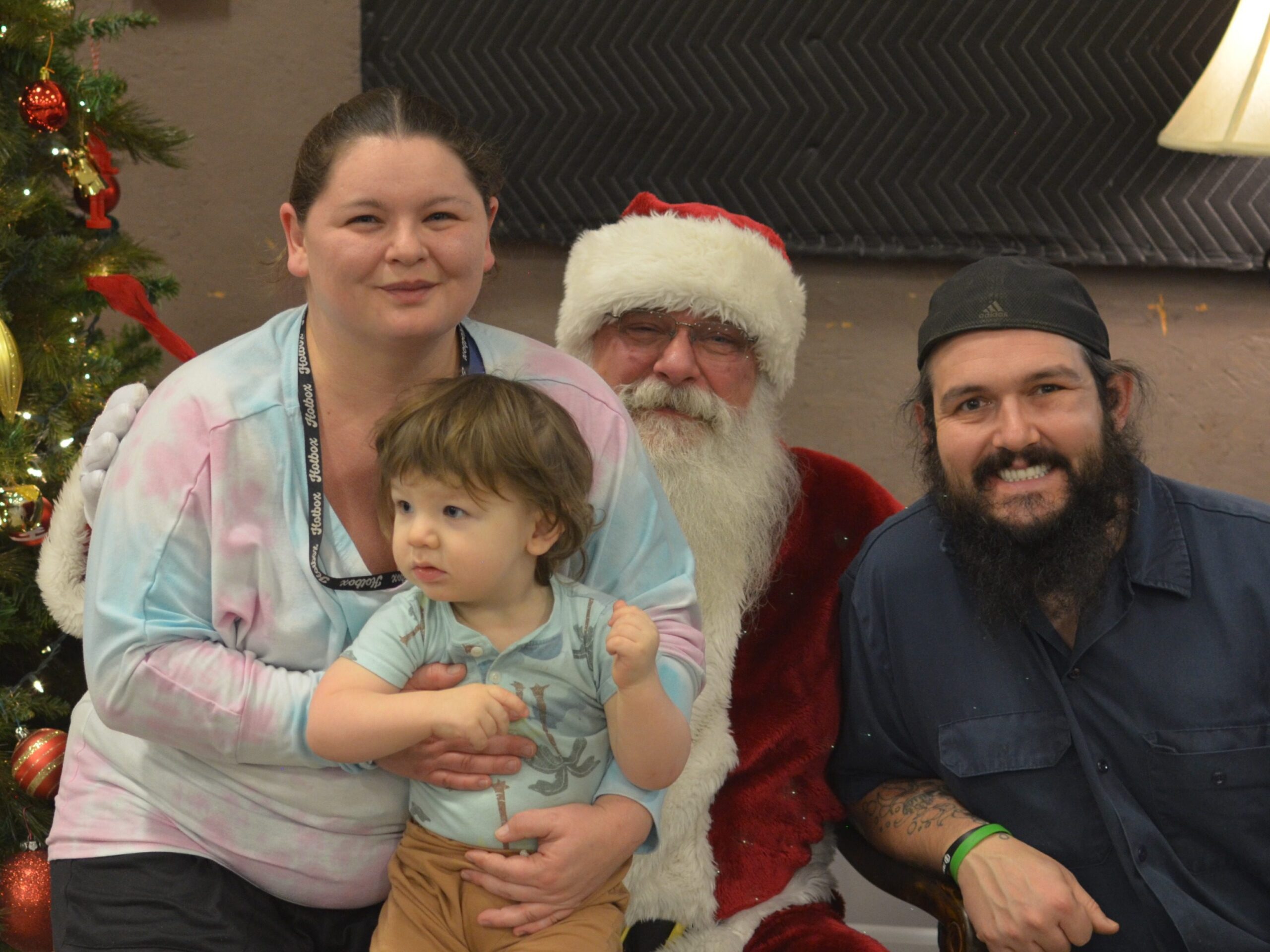 Family posing with Santa Tom beside a decorated Christmas tree during a holiday event.
