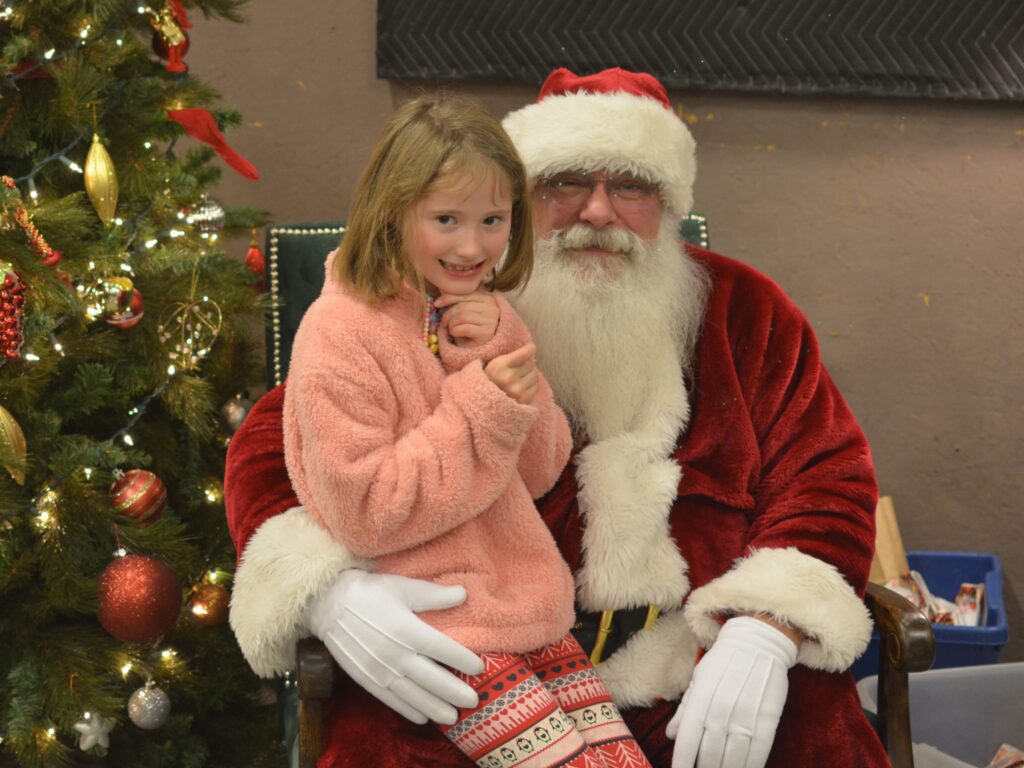 Young girl visiting with Santa Tom beside a decorated Christmas tree.