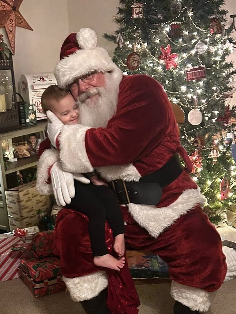 Santa Tom hugging a smiling child beside a Christmas tree during a private family Santa visit in Central Missouri