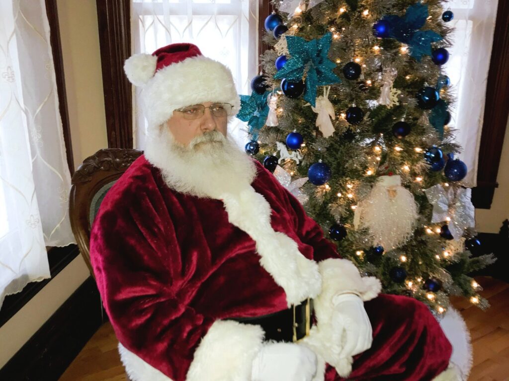 Santa Tom seated beside a decorated Christmas tree