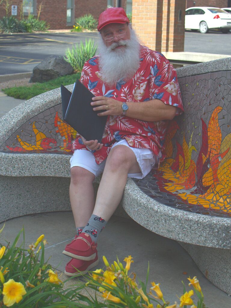 Santa Tom in a red Hawaiian shirt sitting on a decorative bench with a folder in Columbia, Missouri.