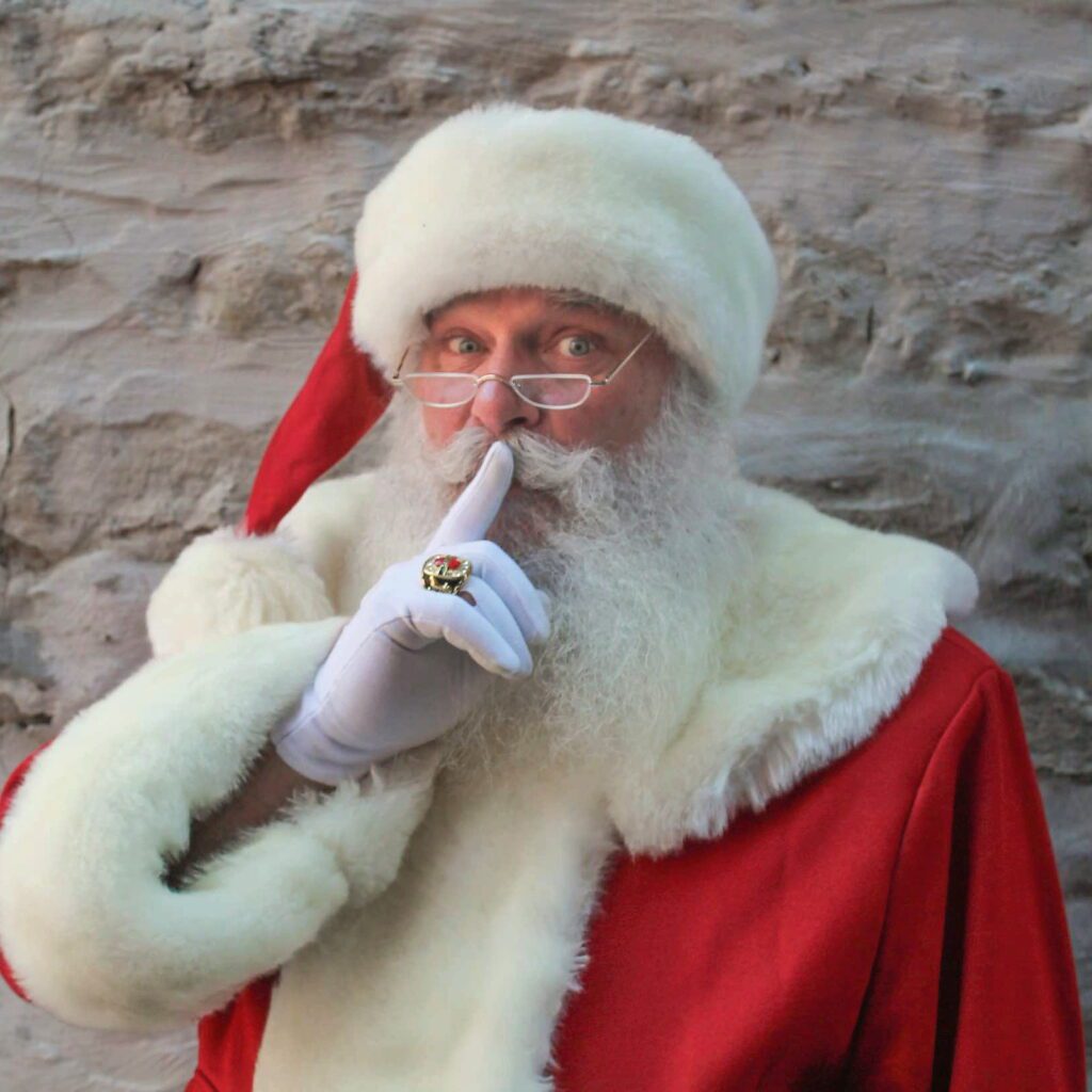 Santa Tom smiling and making a playful shhh gesture.